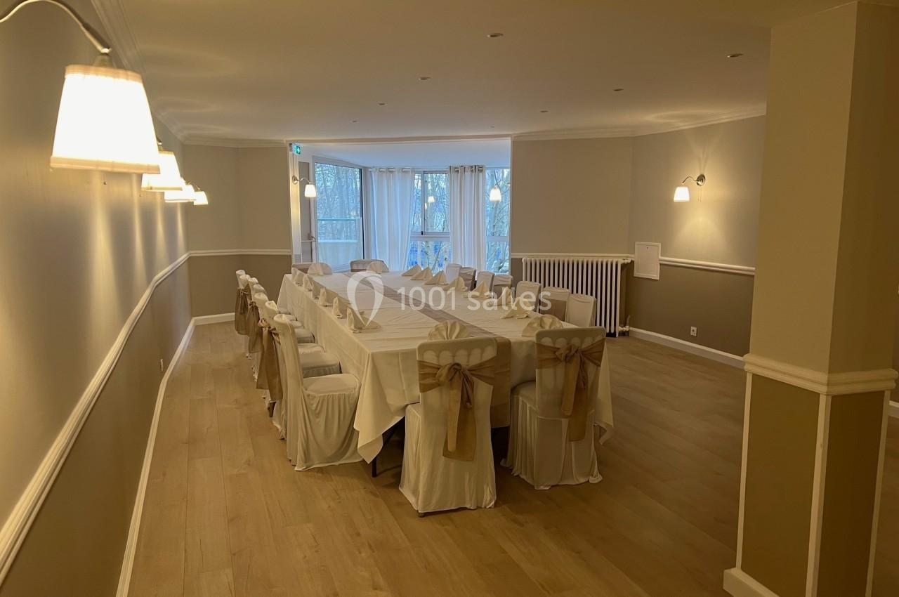 Salle de réception lumineuse avec une grande table rectangulaire dressée et des chaises décorées de nœuds beiges.