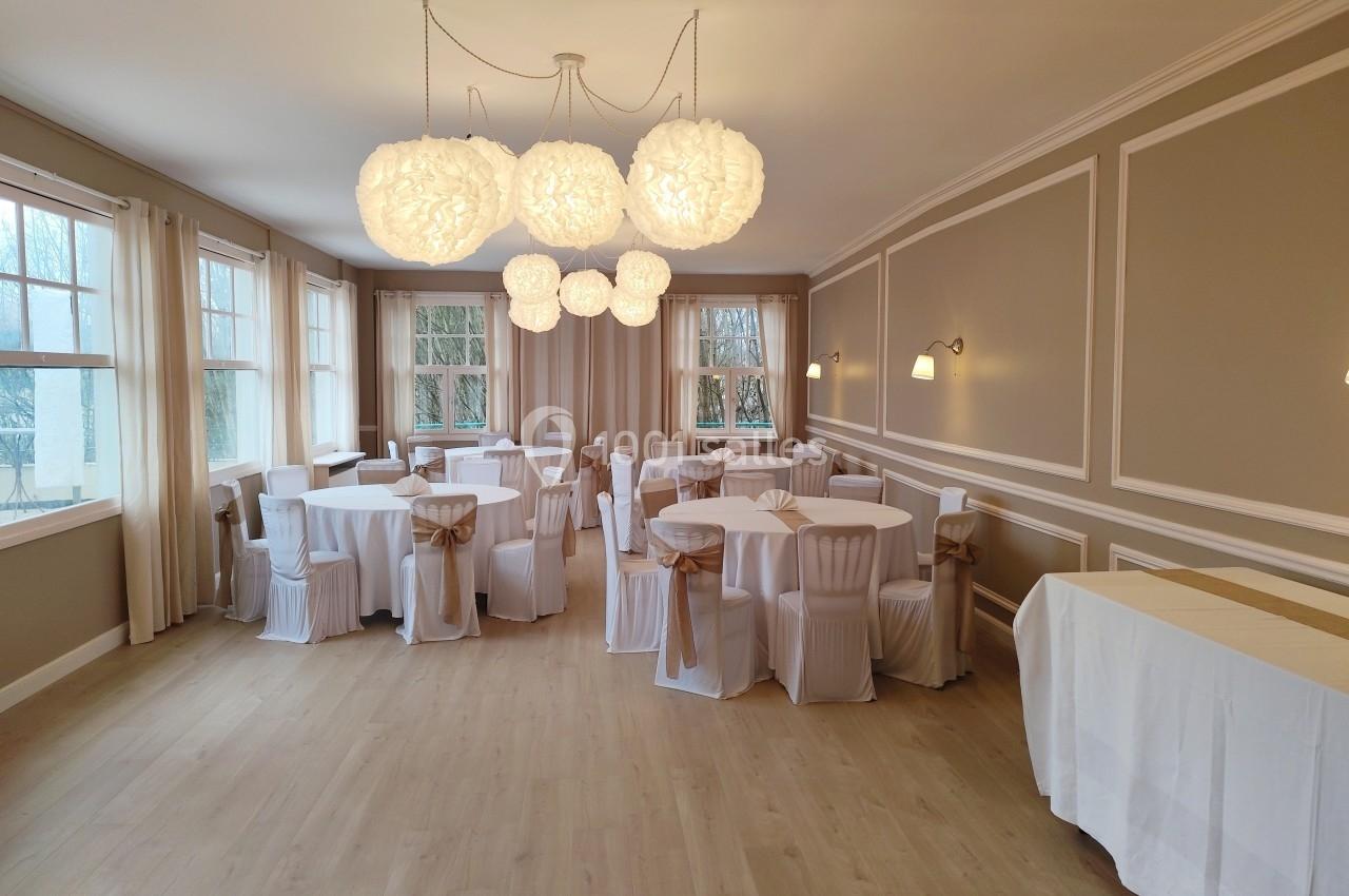 Salle de réception lumineuse avec tables rondes dressées, chaises décorées et suspensions lumineuses.