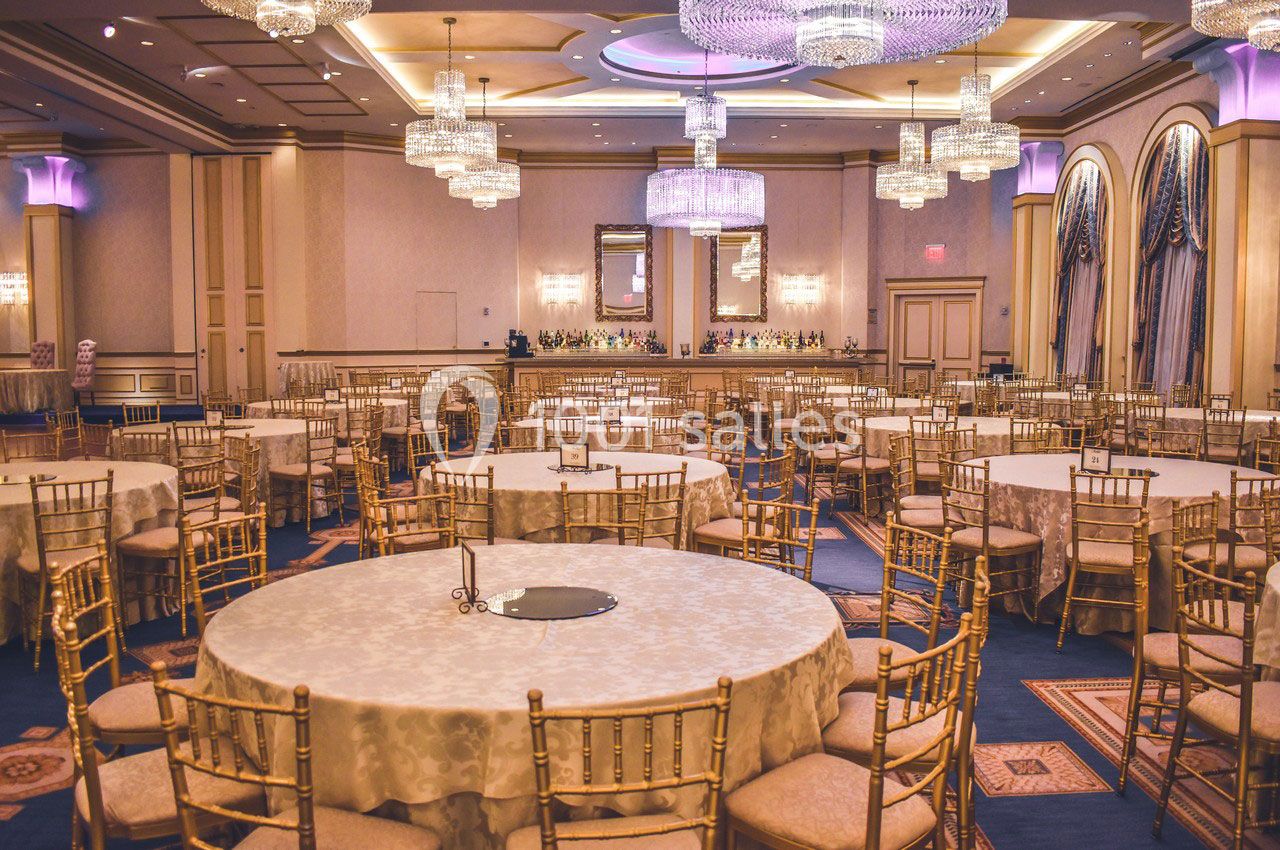 Salle de réception élégante avec tables rondes dressées, chaises dorées et lustres suspendus.