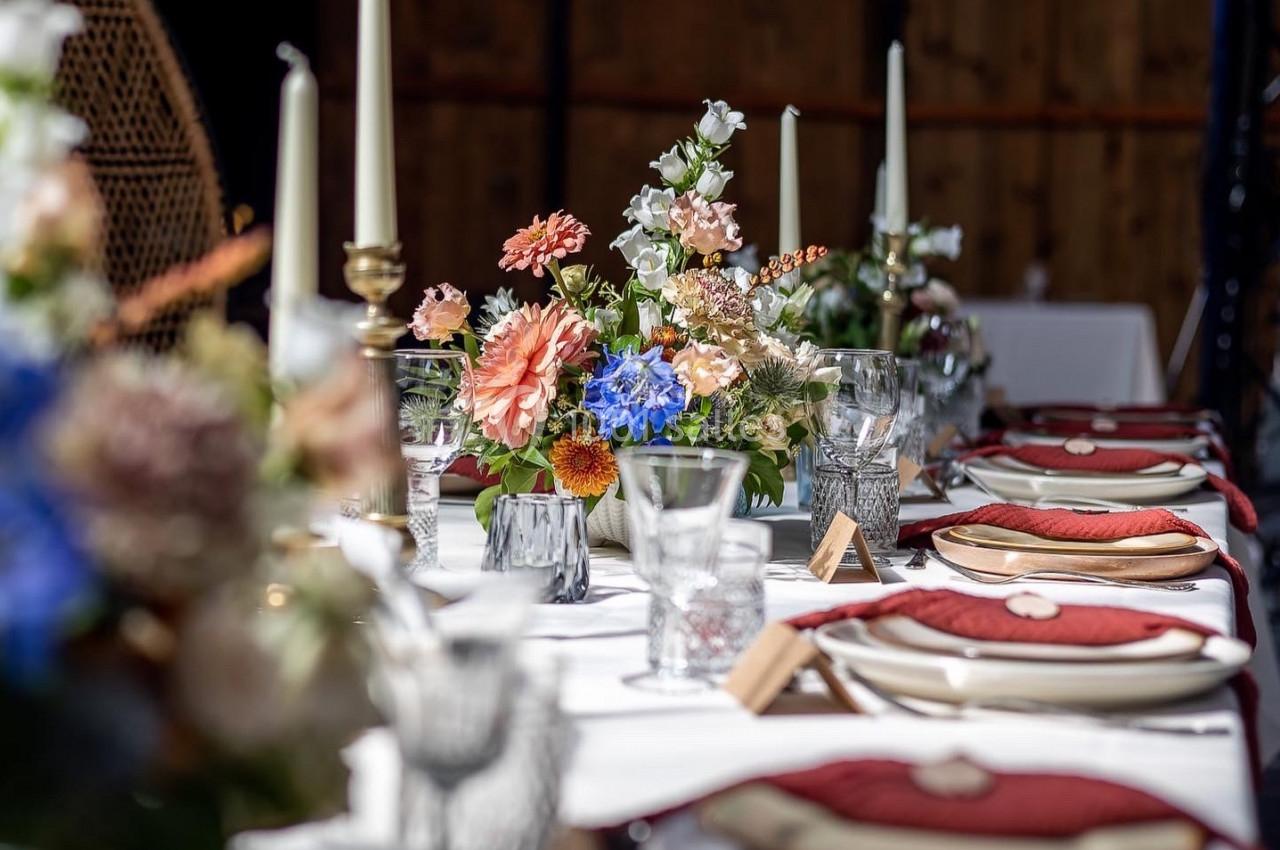 Table dressée avec des assiettes, verres, serviettes rouges et un centre de table floral coloré, éclairée par la lumière…