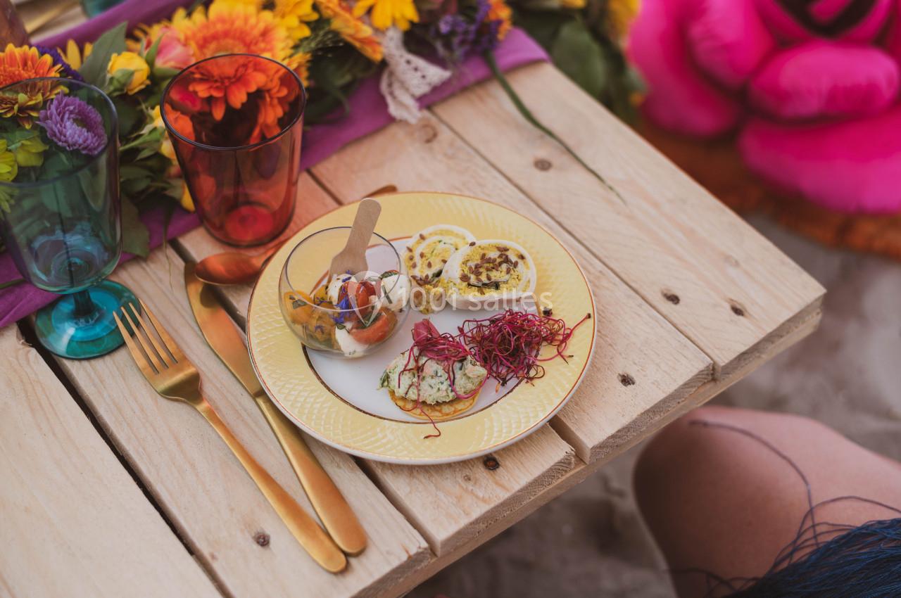 Assiette de tapas colorées sur une table en bois, entourée de fleurs et de verres colorés.