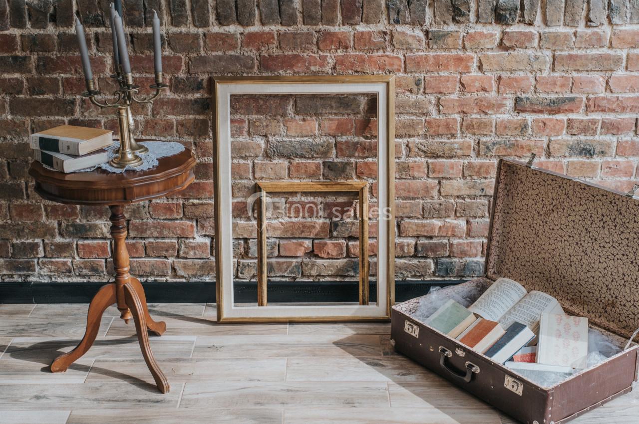 Table en bois avec chandelier, cadres vides appuyés contre un mur de briques et valise ouverte contenant des livres.
