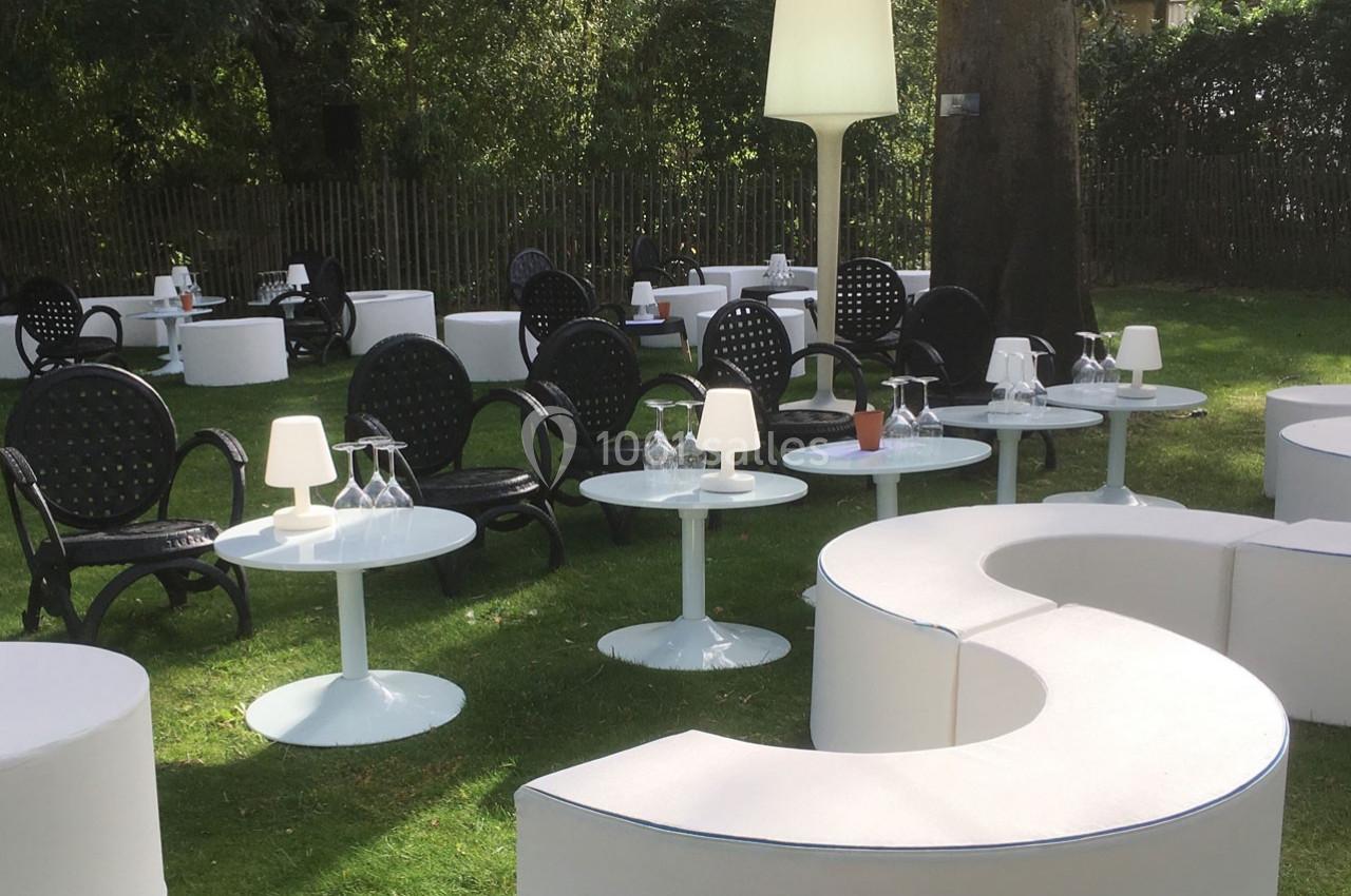 Mobilier de jardin avec tables blanches, chaises noires et lampes, disposé sur une pelouse verdoyante près d'arbres.