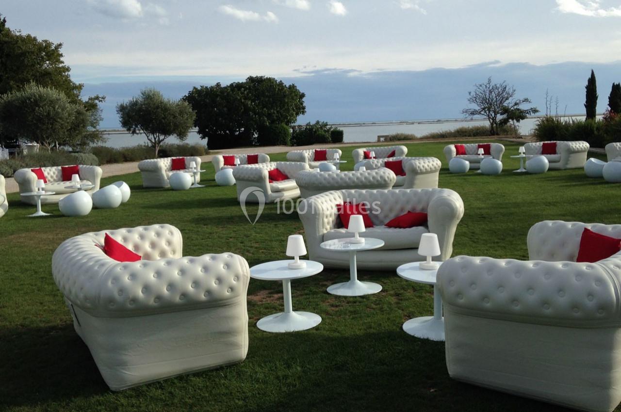 Mobilier de salon blanc avec coussins rouges disposé sur une pelouse en extérieur, près d'un plan d'eau.