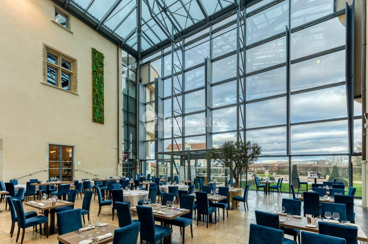 Salle de restaurant lumineuse avec de grandes baies vitrées, des tables dressées et des chaises bleues.