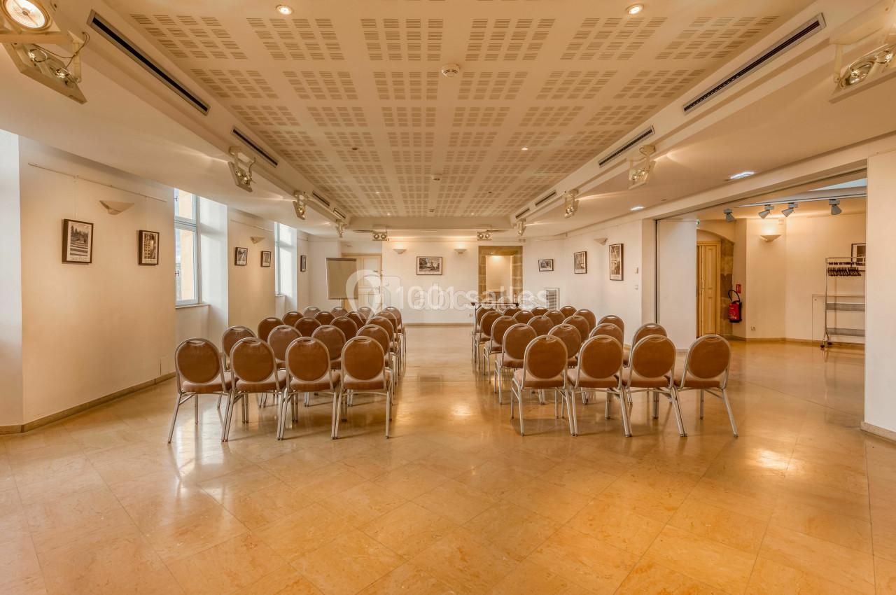 Salle lumineuse avec des rangées de chaises disposées face à un mur blanc, éclairée par des spots au plafond.