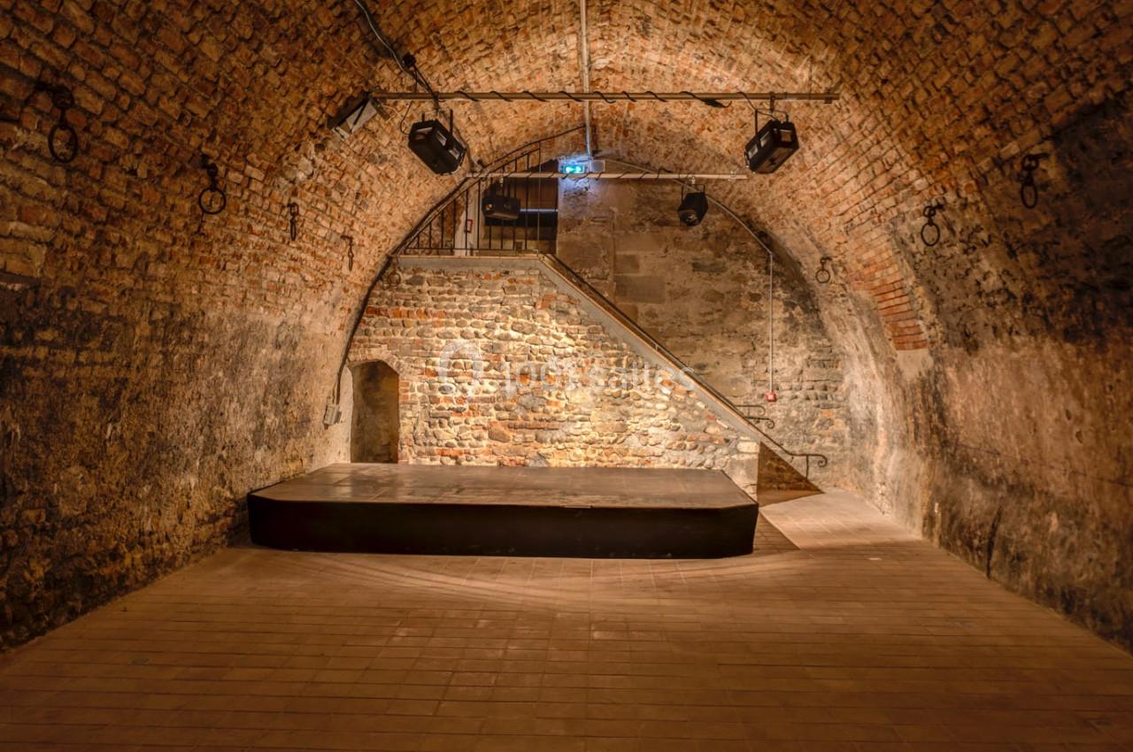 Salle voûtée en briques avec une petite scène surélevée, éclairée par des projecteurs suspendus.