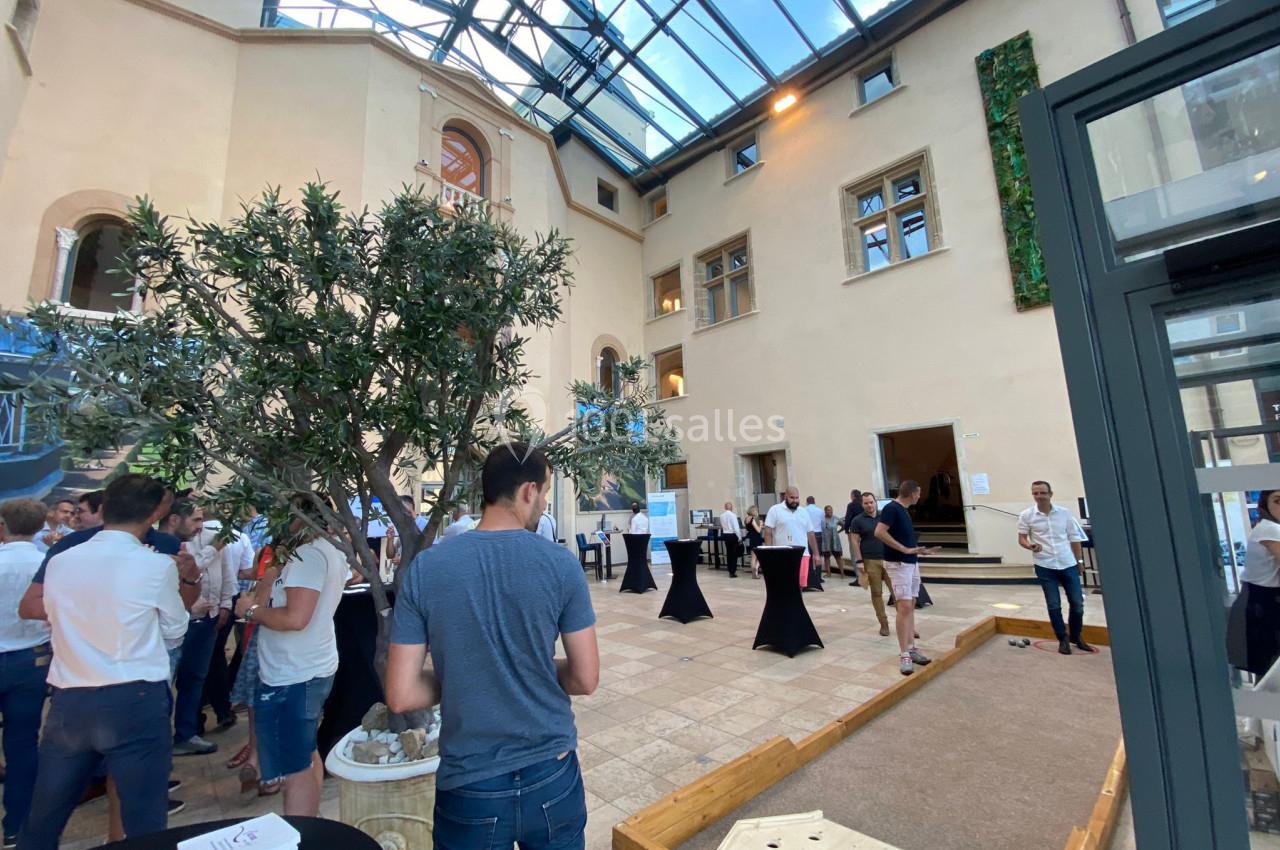 Personnes rassemblées dans une cour intérieure lumineuse avec un toit en verre, un terrain de pétanque et des tables hautes.