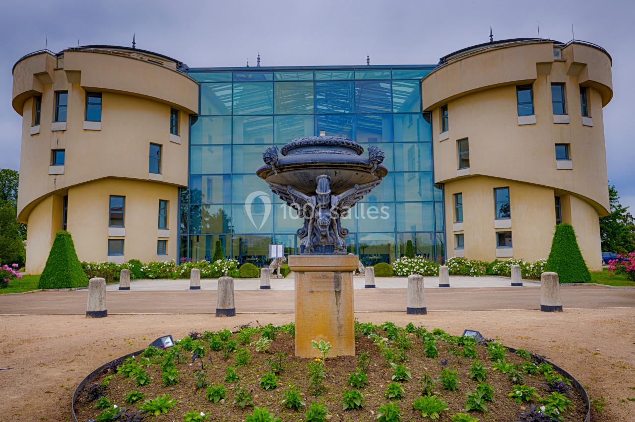 Façade moderne en verre entourée de deux bâtiments arrondis, avec une fontaine et des parterres fleuris au premier plan.