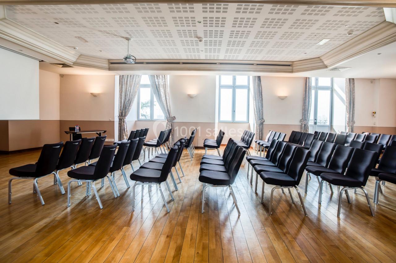 Salle lumineuse avec parquet, rangées de chaises noires alignées face à un écran de projection.