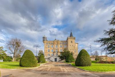 Façade d'un château historique entouré de pelouses bien entretenues et d'arbres taillés, sous un ciel nuageux.