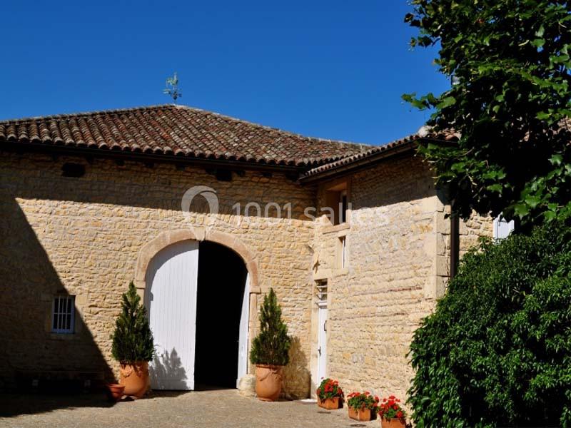 Bâtisse en pierre avec toit en tuiles, grande porte blanche ouverte, pots de fleurs et végétation environnante.
