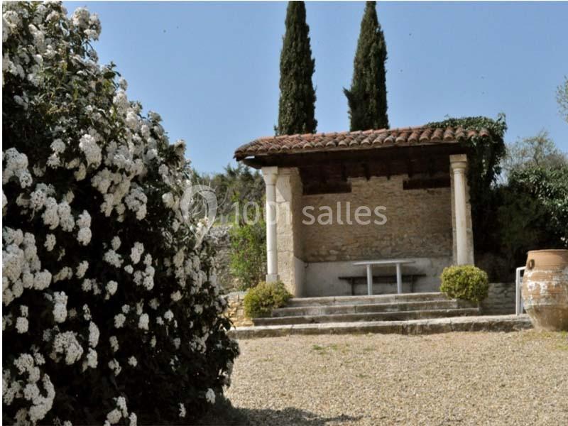 Abri en pierre avec toit en tuiles, entouré de cyprès et de fleurs blanches, sur une cour gravillonnée.