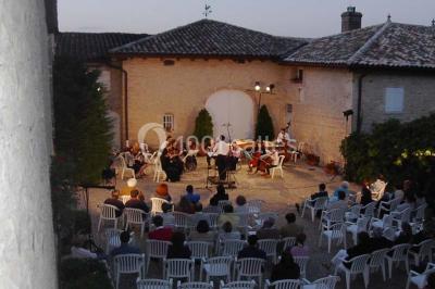 Concert en plein air dans une cour pavée au crépuscule, avec un orchestre et des spectateurs assis sur des chaises blanches.