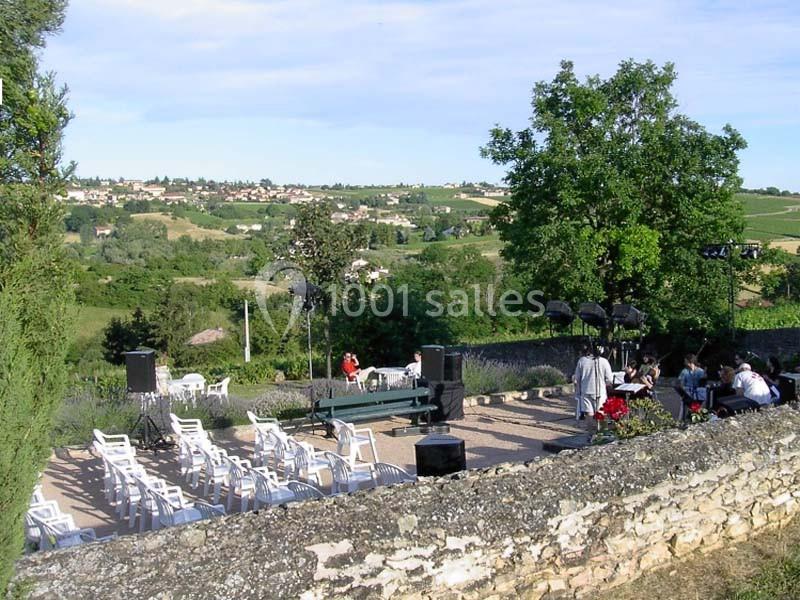 Terrasse en plein air avec des chaises blanches disposées face à une scène, vue sur une campagne vallonnée.