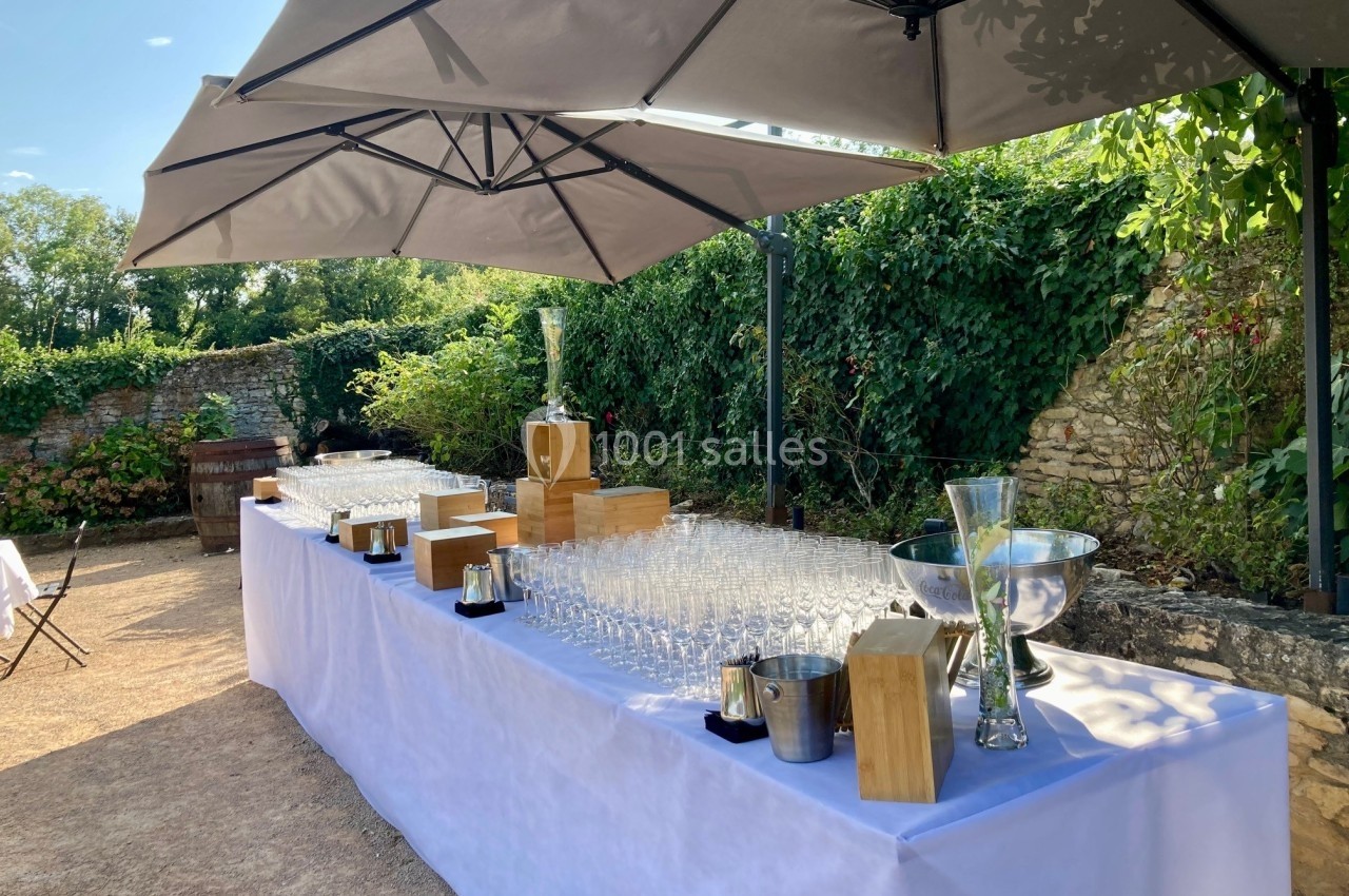 Table dressée en extérieur avec des verres, seaux à glace et décorations sous des parasols, entourée de verdure.