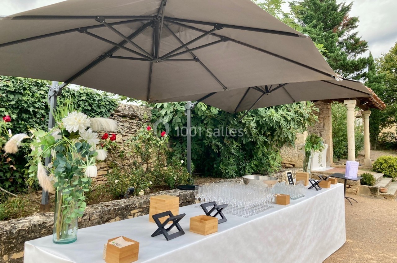 Table dressée en extérieur avec nappage blanc, verres alignés, décorations florales et parasols gris.