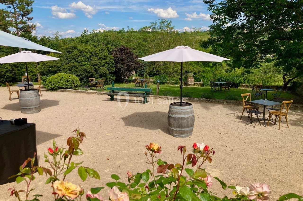 Terrasse extérieure avec tables et parasols sur un sol en gravier, entourée de verdure et de fleurs au premier plan.