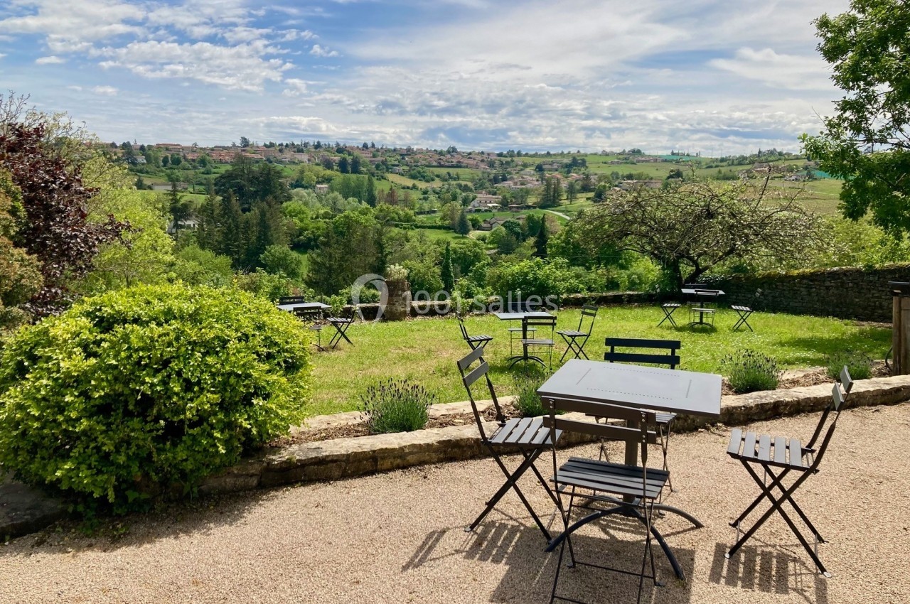 Terrasse avec tables et chaises en métal noir, offrant une vue sur un paysage vallonné et verdoyant sous un ciel…