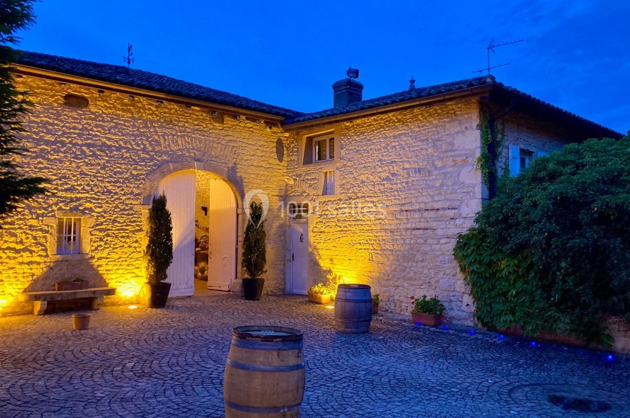 Cour pavée éclairée au crépuscule, entourée de murs en pierre, avec des tonneaux et des plantes décoratives.