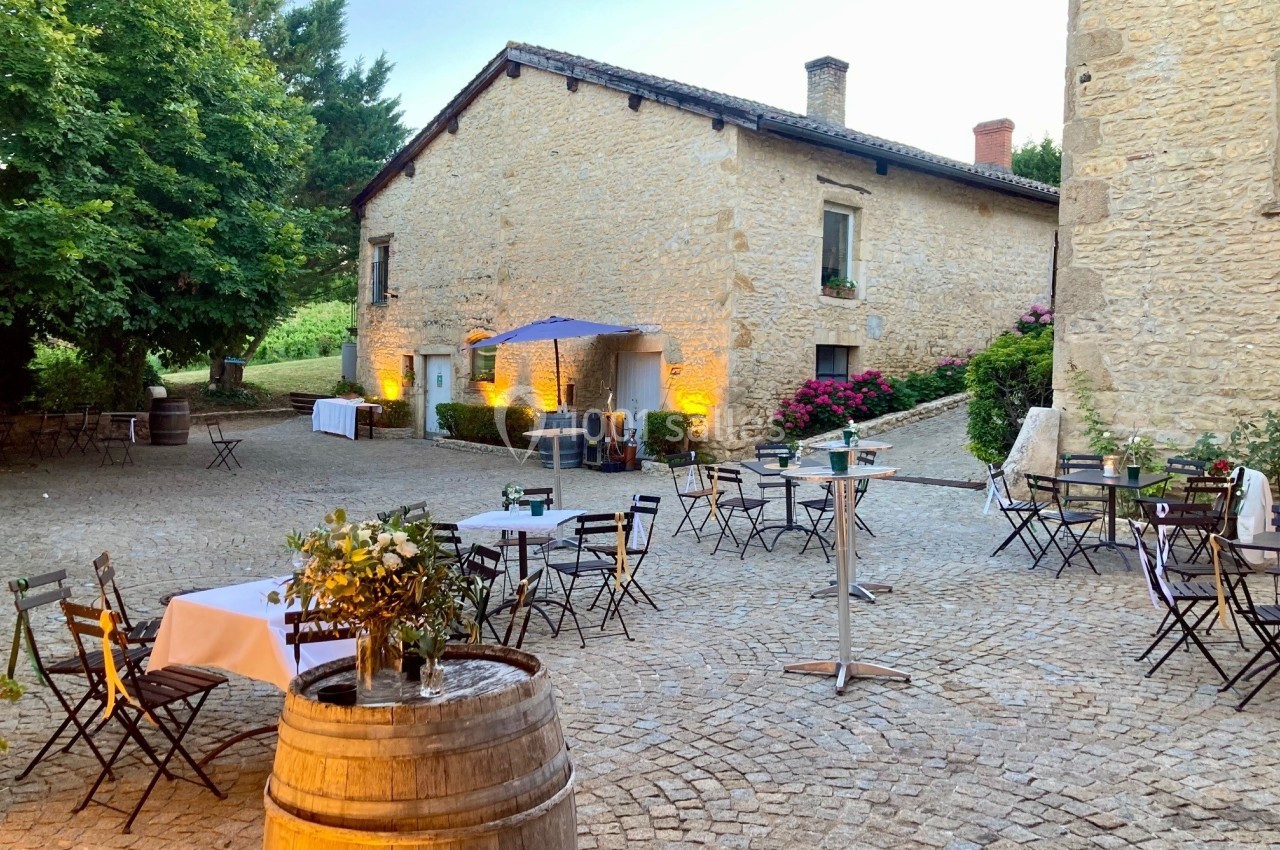Cour pavée avec tables et chaises disposées autour d'un tonneau, entourée de bâtiments en pierre et de végétation.