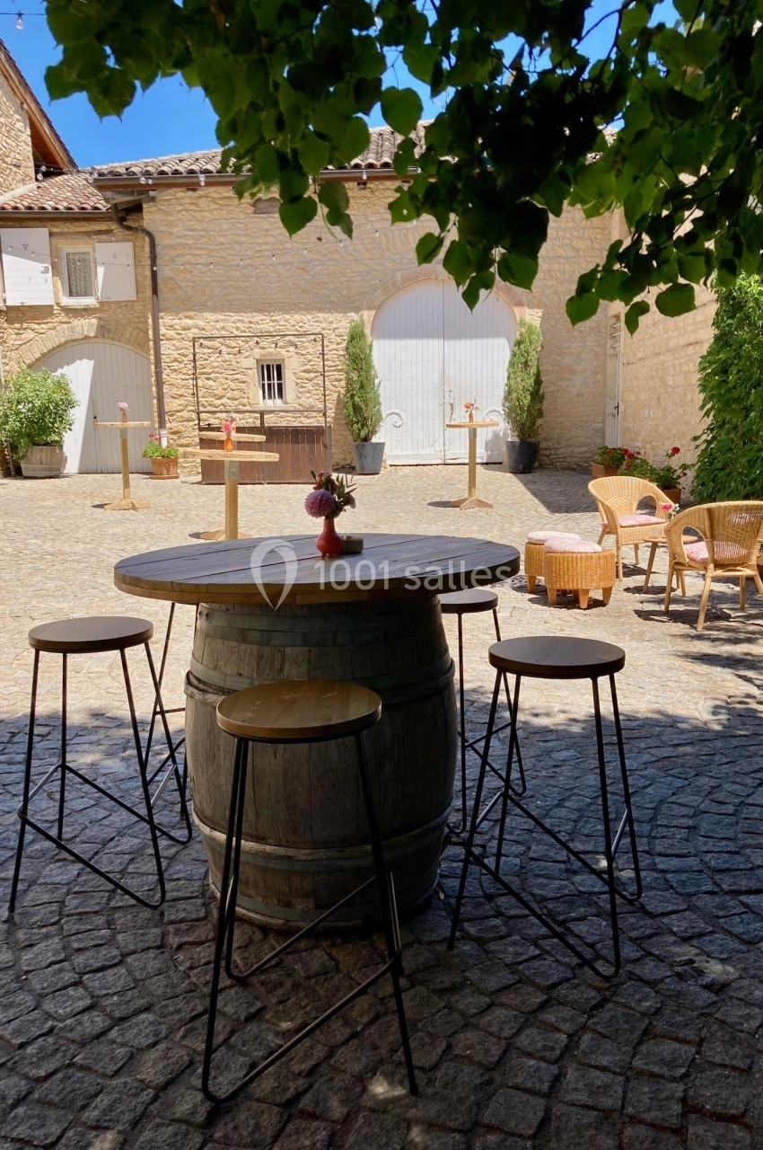 Table haute en tonneau entourée de tabourets sur une cour pavée, avec des bâtiments en pierre en arrière-plan.