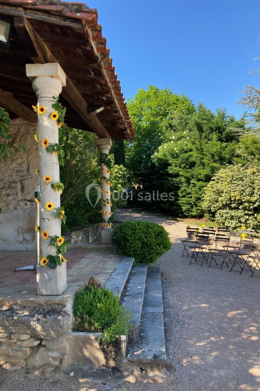Terrasse en pierre avec colonnes décorées de tournesols, entourée de verdure, et chaises disposées en extérieur.