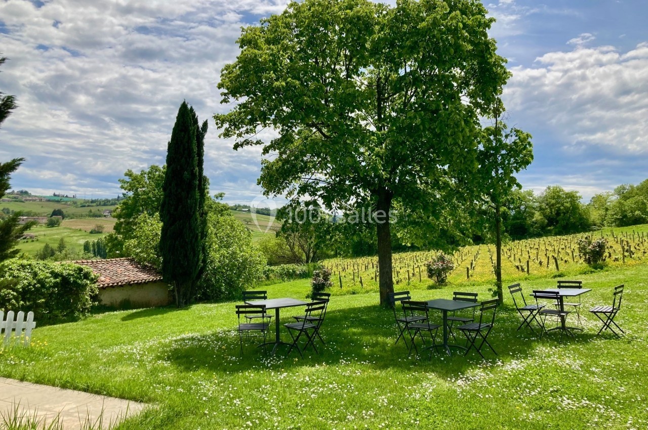 Tables et chaises en métal disposées sur une pelouse verdoyante sous des arbres, avec un paysage rural en arrière-plan.