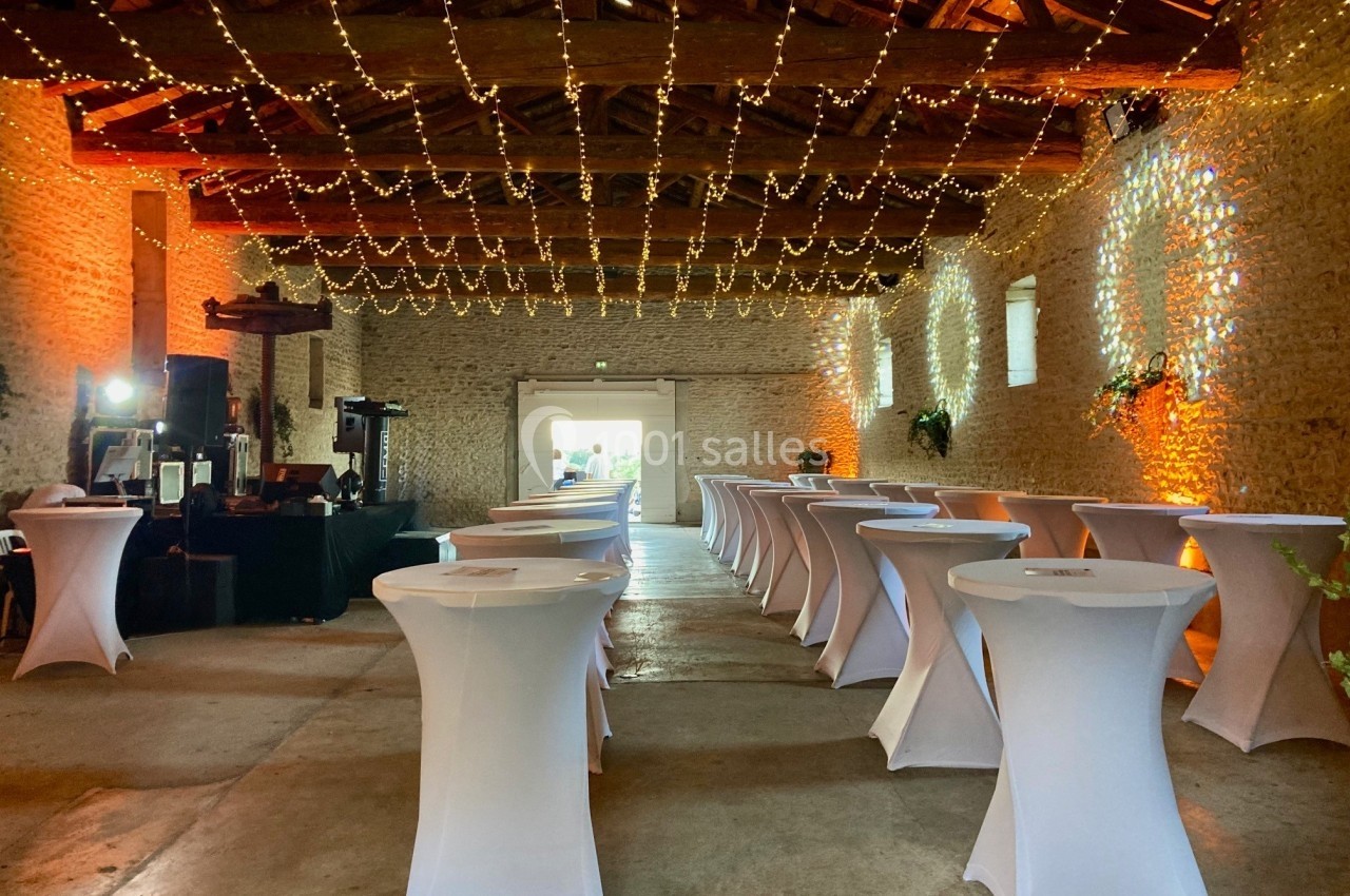 Grande salle en pierre avec tables hautes recouvertes de nappes blanches, éclairée par des guirlandes lumineuses suspendues.