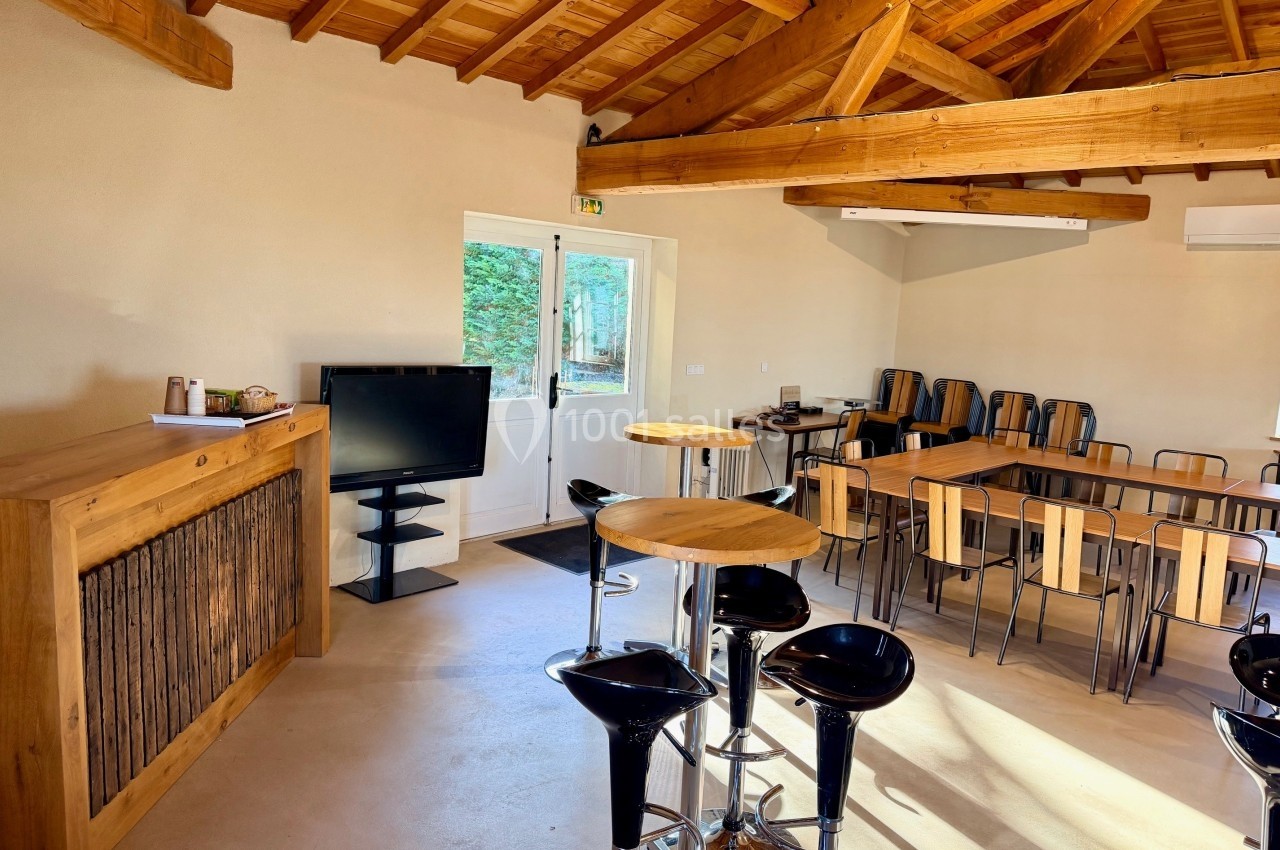 Salle lumineuse avec poutres apparentes, tables en bois, chaises, tabourets hauts et écran près d'une porte vitrée.