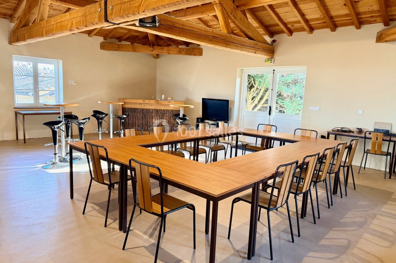 Salle lumineuse avec charpente en bois, tables disposées en U, chaises, bar en fond et porte ouverte sur l'extérieur.