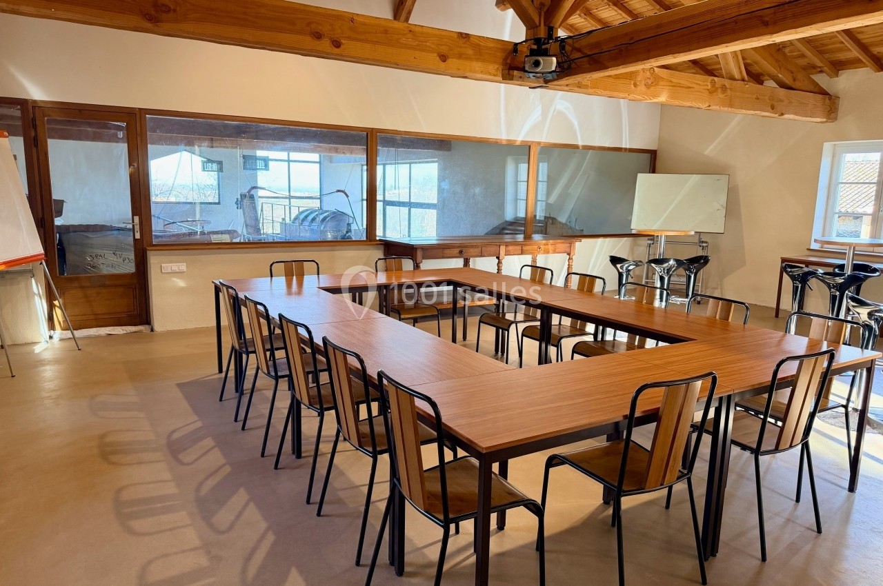 Salle de réunion lumineuse avec tables en U, chaises en bois et vue sur une pièce adjacente à travers une baie vitrée.