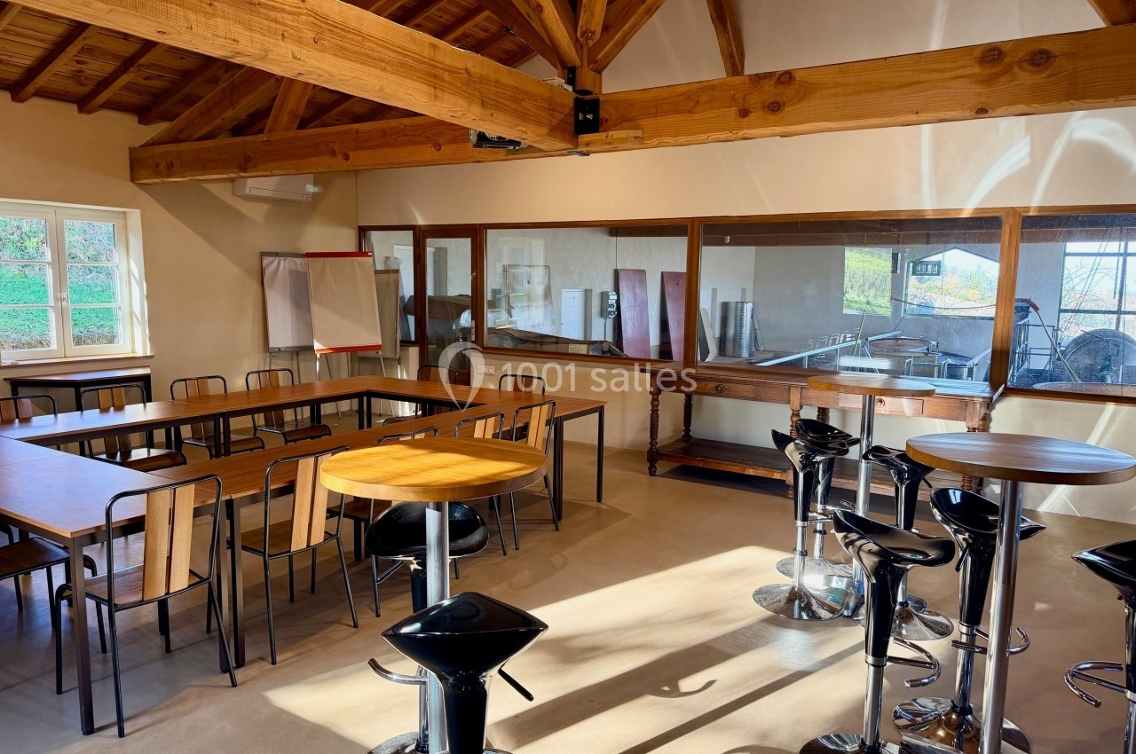 Salle lumineuse avec tables en bois, chaises, tabourets hauts et vue sur un espace vitré donnant sur des équipements.
