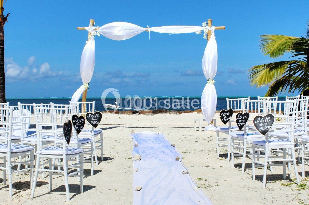 Chaises blanches disposées face à une arche décorée de voiles blancs sur une plage avec vue sur la mer.
