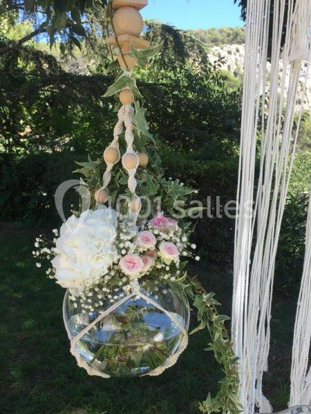 Vase suspendu en macramé contenant des fleurs blanches et roses, décoré de feuillage, dans un jardin verdoyant.