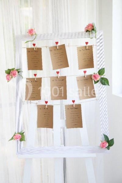 Tableau blanc décoré de fleurs roses, affichant des cartes kraft suspendues avec des pinces en forme de cœur.