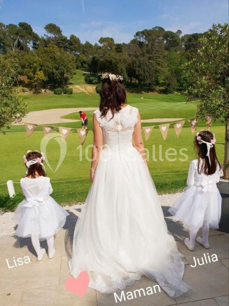 Une femme en robe de mariée de dos, accompagnée de deux petites filles en robes blanches, devant un espace vert.