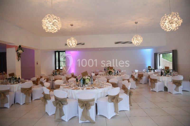 Salle de réception décorée avec des tables rondes, nappes blanches, chaises ornées de nœuds et éclairage suspendu.