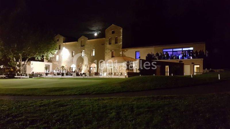 Bâtiment éclairé de nuit avec une verrière et des fenêtres illuminées, entouré de pelouse et d'arbres.