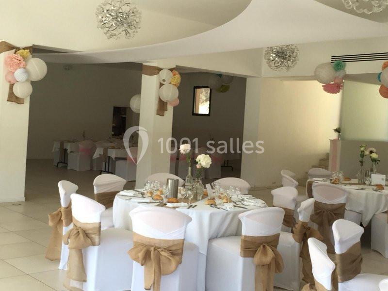 Salle de réception décorée avec des tables rondes dressées, chaises ornées de nœuds beige et suspensions colorées.
