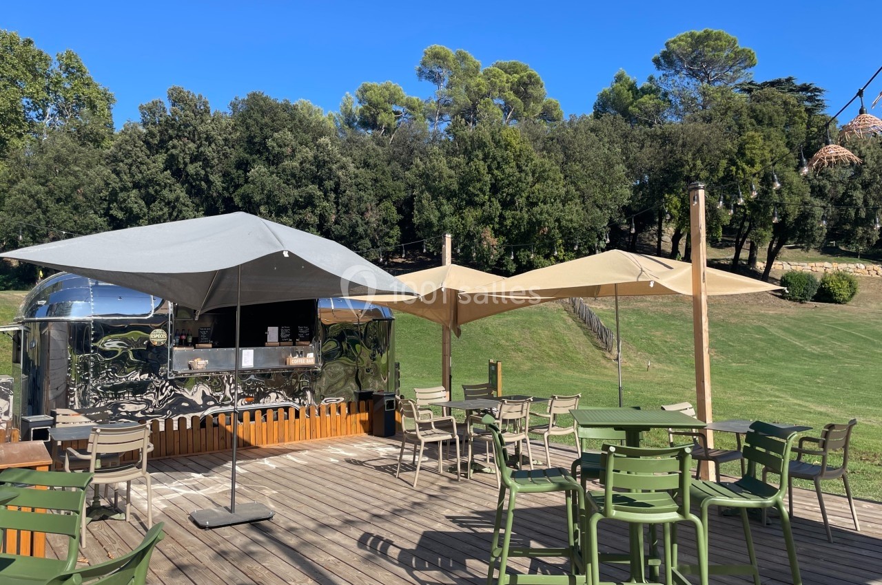 Terrasse en bois avec des tables, des chaises, des parasols et un food truck argenté, entourée de verdure.