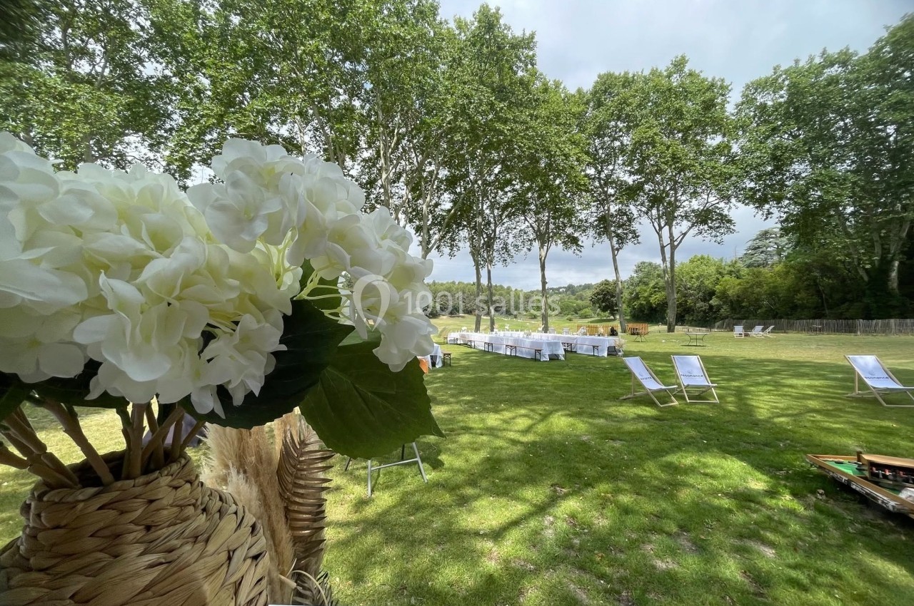 Jardin verdoyant avec des tables dressées et des transats sous des arbres, vue partielle d'un bouquet de fleurs blanches.