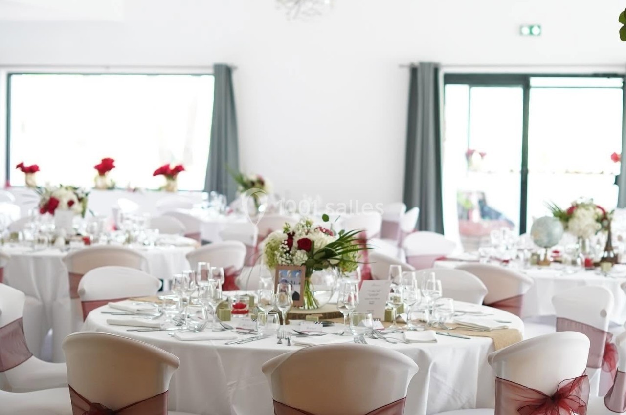 Salle de réception lumineuse avec tables rondes décorées de nappes blanches, rubans rouges et bouquets de fleurs.