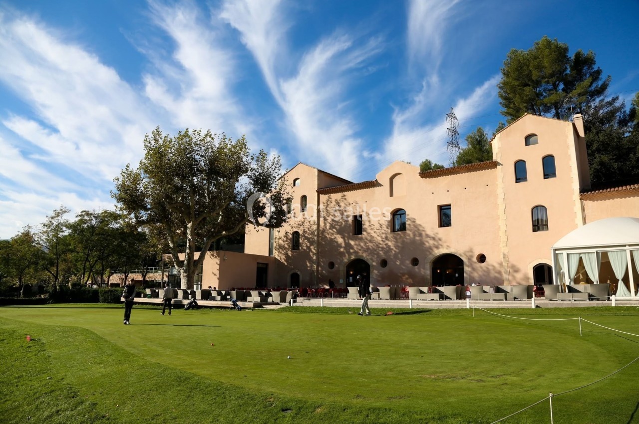 Bâtiment beige entouré d'arbres avec un terrain de golf au premier plan sous un ciel bleu parsemé de nuages.