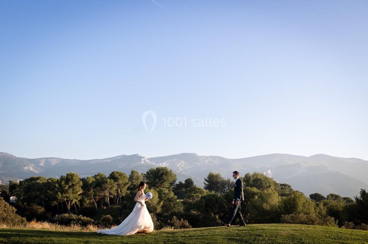 Une mariée en robe blanche marche sur une pelouse, suivie par un homme en costume, avec des montagnes en arrière-plan.