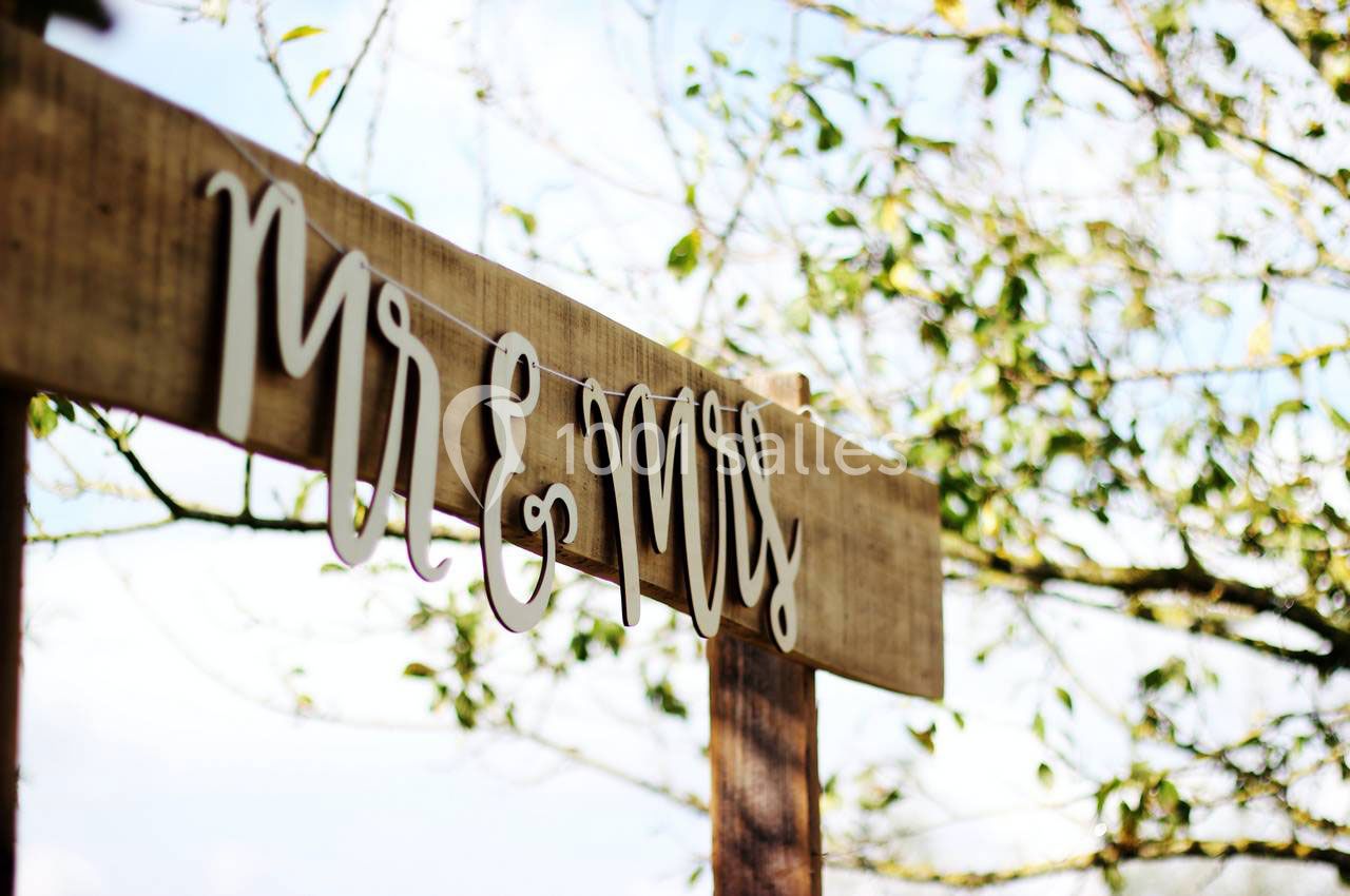 Panneau en bois avec l'inscription ’Mr & Mrs’ suspendue, devant des branches d'arbres et un ciel partiellement dégagé.