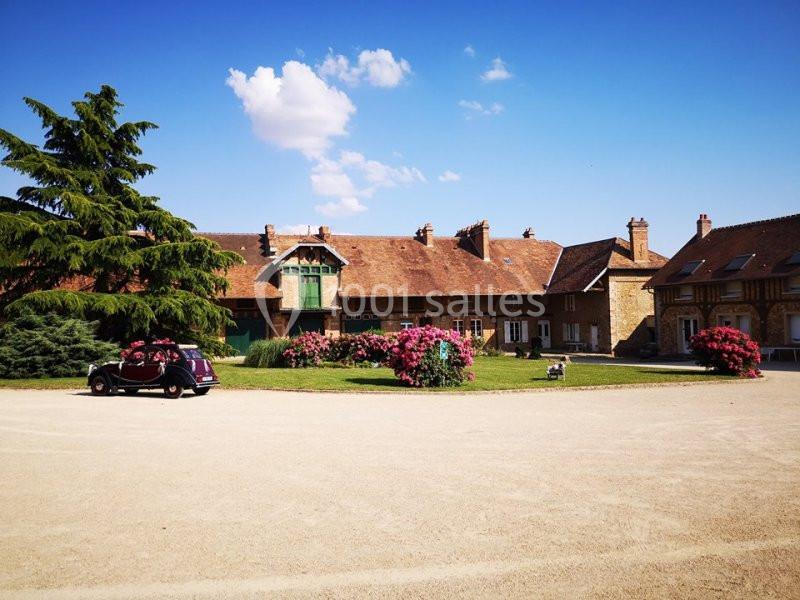 Cour d'une grande maison en pierre avec des fleurs colorées, une voiture ancienne et un ciel bleu dégagé.