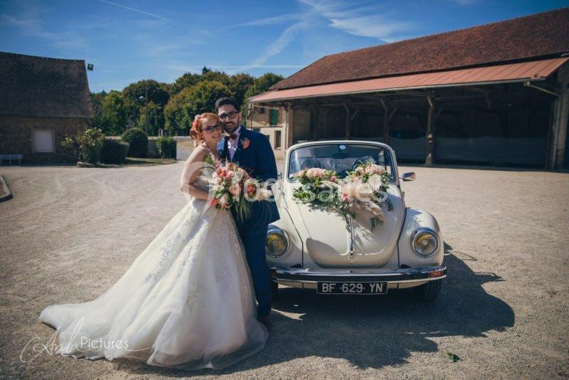 Un couple en tenue de mariage pose devant une voiture ancienne décorée de fleurs, dans une cour ensoleillée.
