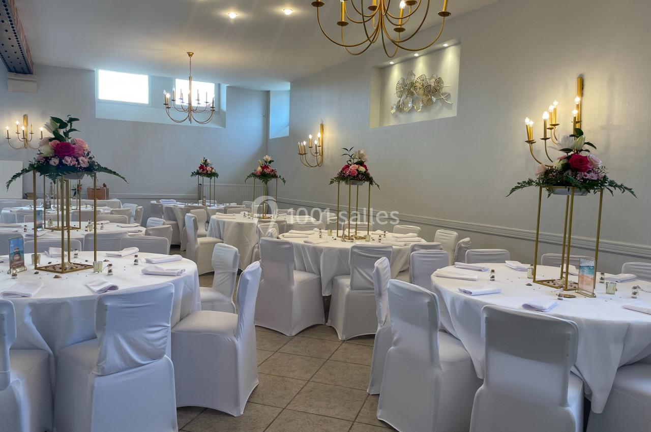 Salle de réception élégante avec tables rondes dressées, nappes blanches et décorations florales sur supports dorés.