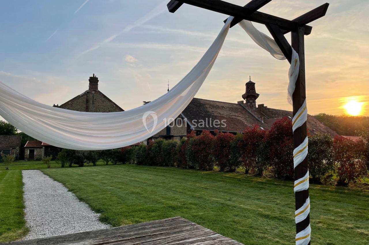 Voile blanc flottant sur une pergola en bois, avec des bâtiments anciens et un coucher de soleil en arrière-plan.